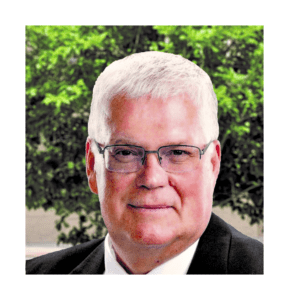 A man with white hair and glasses in front of trees.