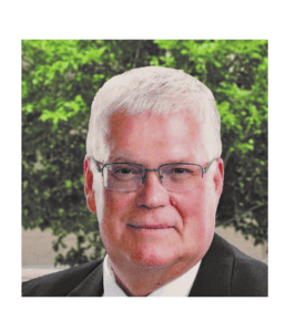 A man with white hair and glasses in front of trees.