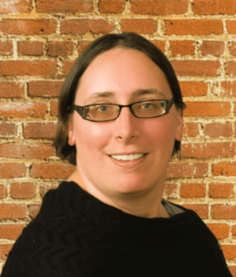 A woman with glasses standing in front of a brick wall.